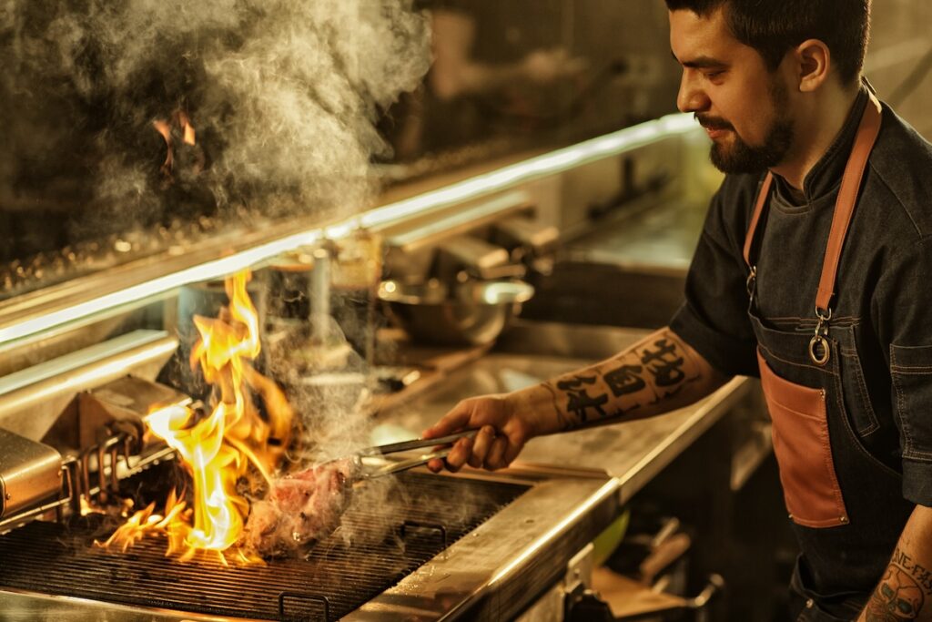 Side view of professional chef cooking delicious juicy beef steak on flaming grill. Handsome man with beard and tattoos on hand preparing food in modern restaurant kitchen.